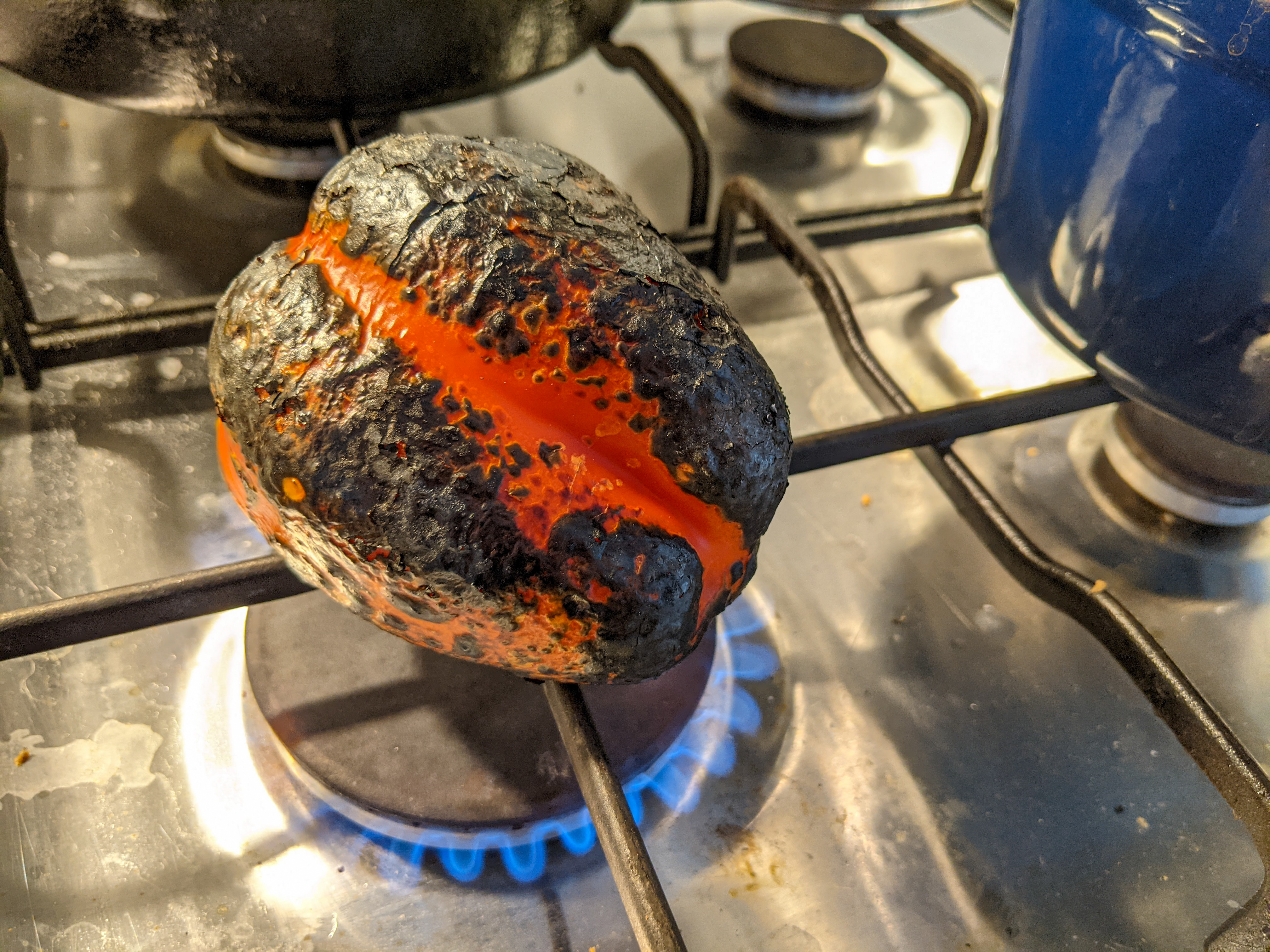 A red pepper sitting on the lit burner of a gas stove. It has significant blackening on the surface